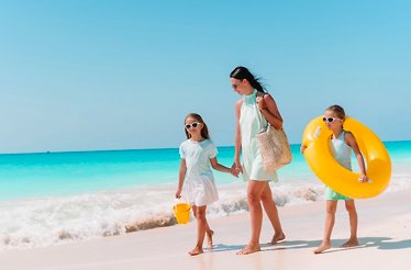 Mother and two young girls walking along a sandy beach, one child carrying a yellow inflatable ring and bucket.