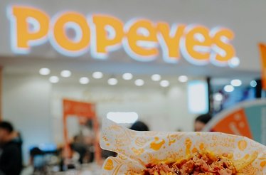 Popeyes branded food tray held in front of a blurred Popeyes restaurant storefront with illuminated yellow signage.