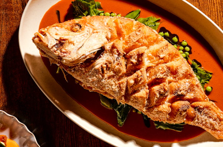 Salt-crusted whole roasted fish plated over vibrant red sauce with snap peas and greens, served at Little Water.