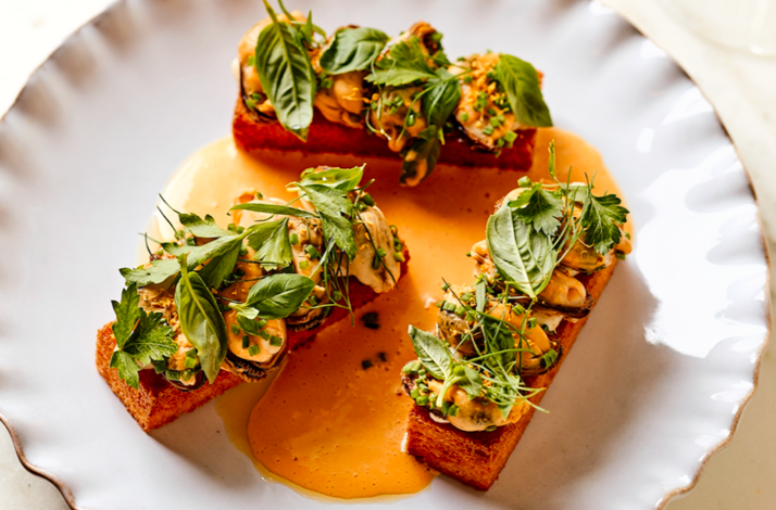 Three toasted brioche pieces topped with clams, fresh herbs, and microgreens in a golden sauce on a scalloped plate at Little Water.