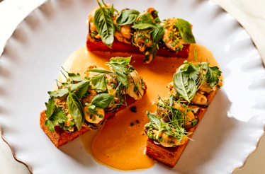 Three toasted brioche pieces topped with clams, fresh herbs, and microgreens in a golden sauce on a scalloped plate at Little Water.