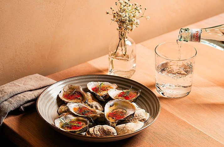 Plate of oysters on the half shell with garnish on a wooden table, beside a glass of water being poured, served at River Twice.