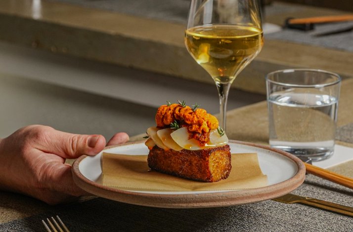 Hand holding a plated toast topped with sliced egg and orange roe, with a glass of white wine and water behind.