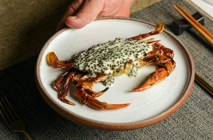 Whole cooked crab served on a ceramic plate, topped with a creamy sauce and small pearls of roe.