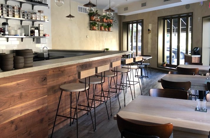 Bright, minimalist restaurant dining room with a long wood bar, stools, and sunlit tables by large windows.