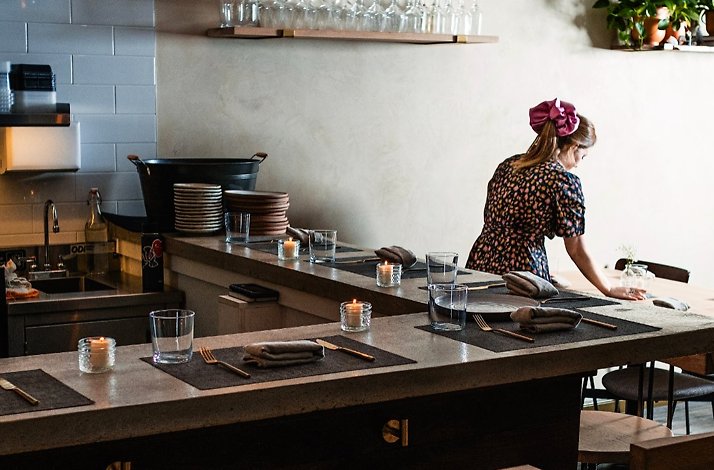 Restaurant interior with counter seating set with candles and place settings, as a staff member arranges a table.