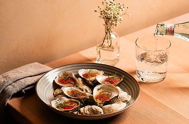 Plate of oysters on the half shell with garnish on a wooden table, beside a glass of water being poured, served at River Twice.