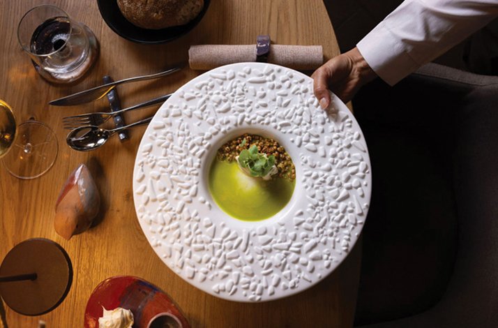 Server placing a sculptured white plate holding a green olive oil-based dish onto a wooden table at Ps Ristorante.