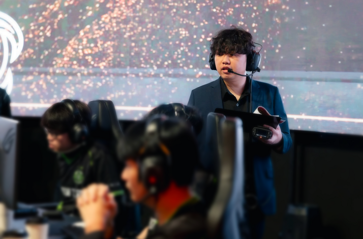 Coach addressing players during a Hong Kong Asian Games Esports Team masterclass, with players at gaming stations on a lit stage.