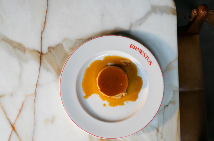 Classic caramel flan topped with amber sauce on an Ernesto's branded white plate, viewed from above.