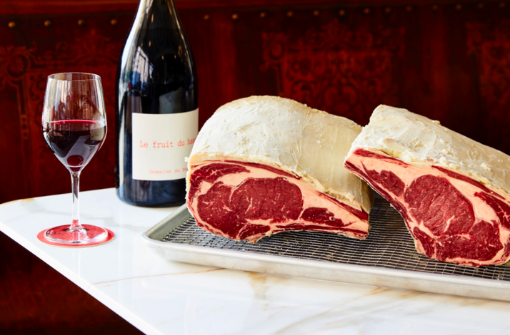 Dry-aged bone-in ribeyes displayed on a wire rack alongside a red wine bottle and glass at Ernesto's. 