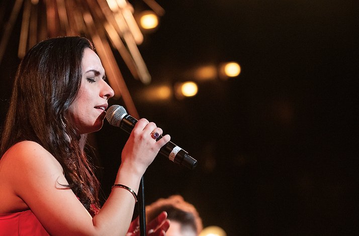 A female vocalist in a red dress is singing into a microphone during a dimly lit live performance.