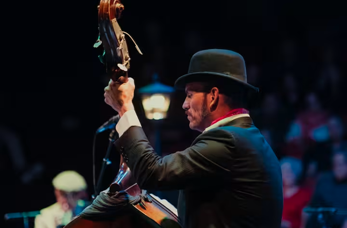 A side profile of a musician in a bowler hat playing a double bass on a dimly lit stage.
