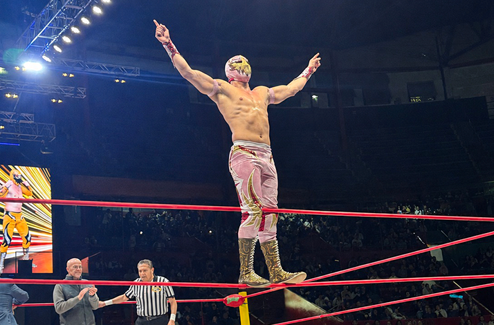 Masked lucha libre wrestler balancing on the corner ropes inside the ring during a live wrestling event illuminated by arena lights.