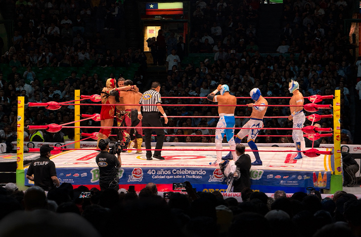 Lucha libre match taking place inside a packed arena, with masked wrestlers competing in the ring as spectators watch from surrounding seats.
