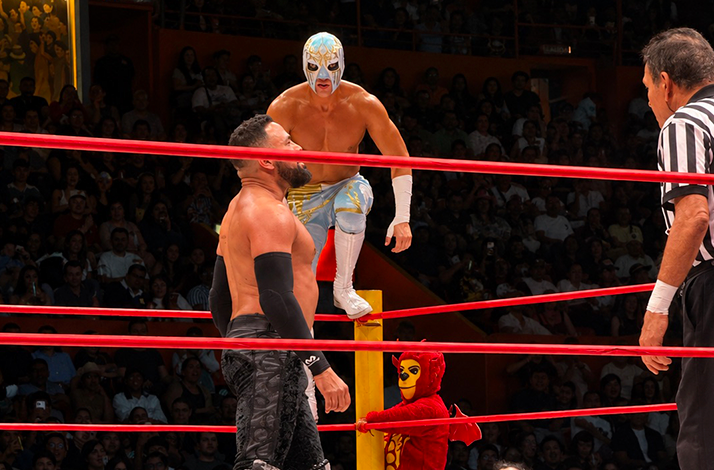 Masked lucha libre wrestlers face off inside the ring during a live match, with spectators surrounding the arena and the referee overseeing the action.