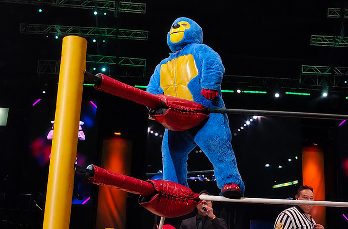 Performer in a colorful costume standing on the ring ropes during a lucha libre event under bright arena lights.
