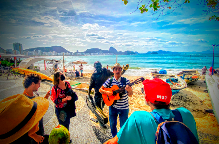 A musician playing guitar along the beachfront in Rio de Janeiro while leading a walking tour, with participants gathered nearby and views of Copacabana Beach and the coastline in the background