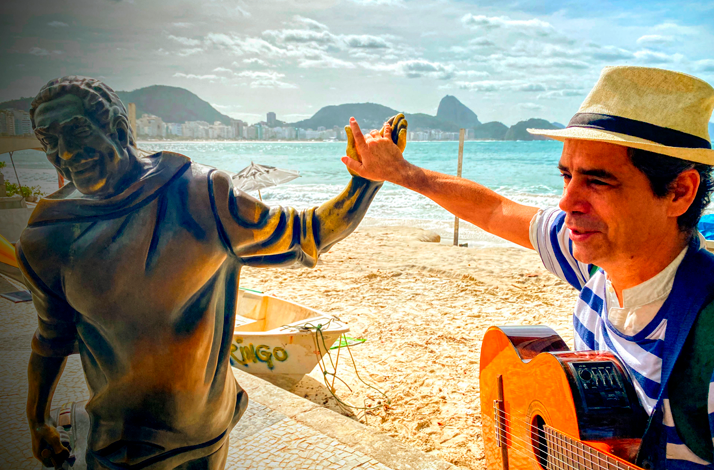 A guitarist standing beside a beachfront statue in Rio de Janeiro, holding a guitar with the ocean, sandy beach, and iconic coastal landscape visible behind