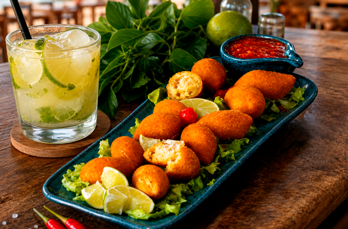 A platter of Brazilian savory snacks served with lime wedges and dipping sauce alongside a lime cocktail on a wooden table.