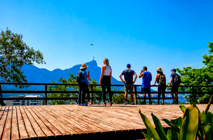 Visitors standing on a scenic observation deck overlooking Rio de Janeiro's mountains and city landscape under a clear blue sky.