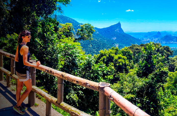 Visitor standing at a scenic lookout in a tropical forest, overlooking Rio de Janeiro's mountains, greenery, and coastline under a bright blue sky.