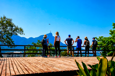 Visitors standing on a scenic observation deck overlooking Rio de Janeiro's mountains and city landscape under a clear blue sky.