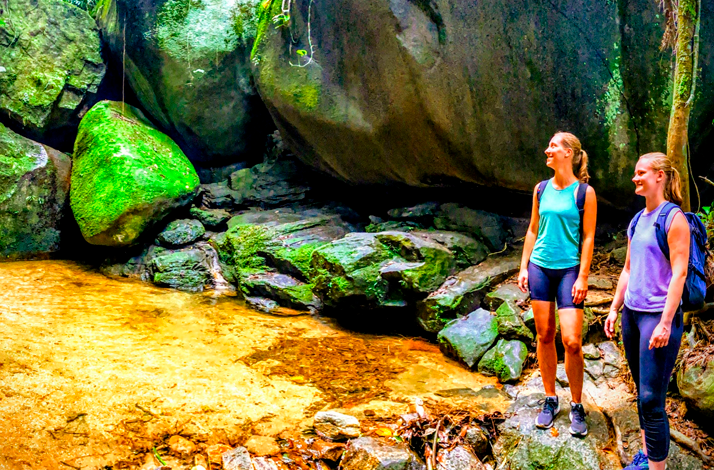 Two hikers explore a rocky jungle stream surrounded by towering moss-covered boulders and lush tropical vegetation in Rio de Janeiro.