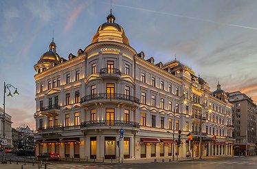 Corinthia Bucharest grand exterior at twilight, showing ornate white facade, domed roofline, wrought-iron balconies, and glowing street-level entrance.