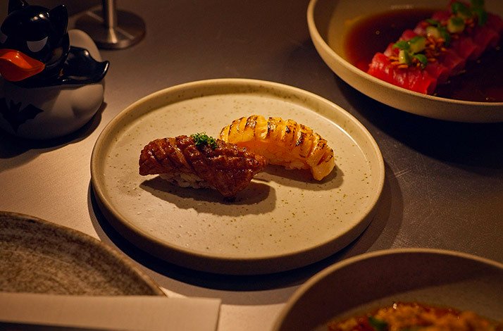 Two scored nigiri pieces on a stone plate next to a Batman duck toy at Kokoro Hand Roll Bar, Dubai.