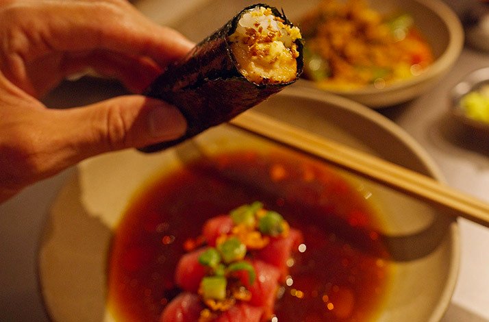 Hand holding a sushi hand roll over a plate of tuna crudo at Kokoro Hand Roll Bar, Dubai.