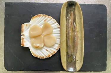 Scallop meats served in a shell alongside a whole anchovy in a glazed dish, plated on slate at O Balado.