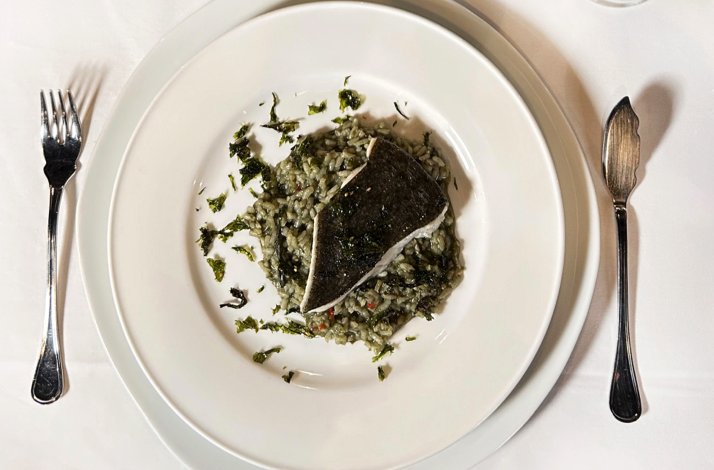 Grilled fish fillet served over dark herb-infused rice on a white plate at Casa Barqueiro restaurant, flanked by fork and knife.