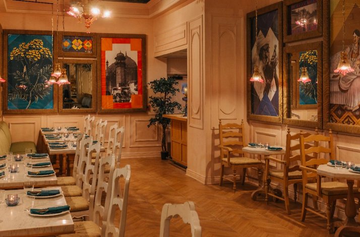 Restaurant interior featuring set dining tables, pendant lights, and colorful Indian-themed paintings on warm walls.