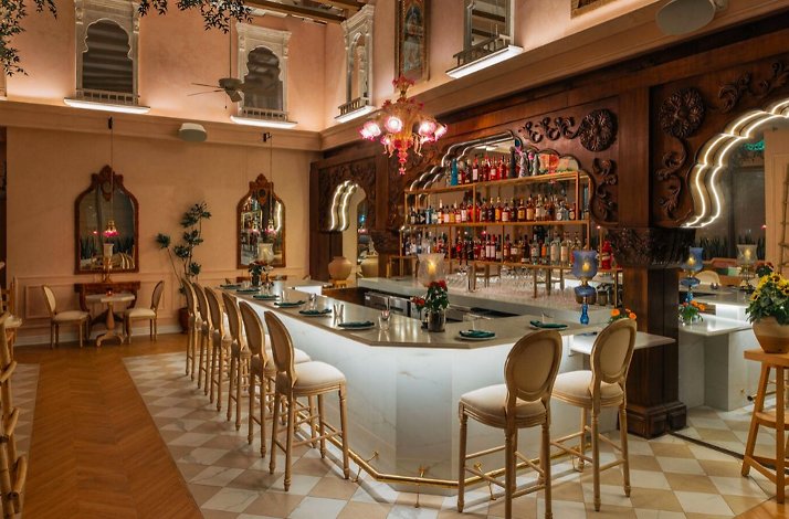 Spacious restaurant bar with a curved marble counter, gold bar stools, carved wood panels, and a pink chandelier overhead.