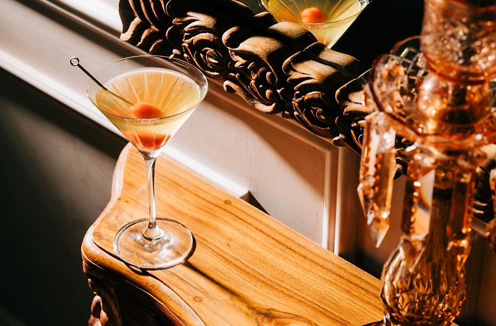 Martini garnished with a cherry and skewer, resting on an ornate wooden surface beside a vintage amber glass candlestick.