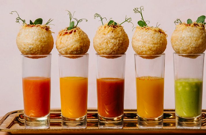 Row of five pani puri garnished with microgreens, each resting on a tall glass of flavored water in varying colors.