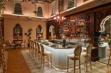 Spacious restaurant bar with a curved marble counter, gold bar stools, carved wood panels, and a pink chandelier overhead.