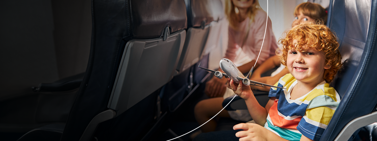 Wide airplane cabin view with a smiling child holding a toy plane in the foreground and family in the background.