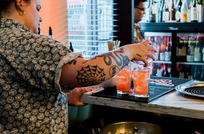 Bartender pours a bright pink cocktail into two ice-filled glasses at the bar.