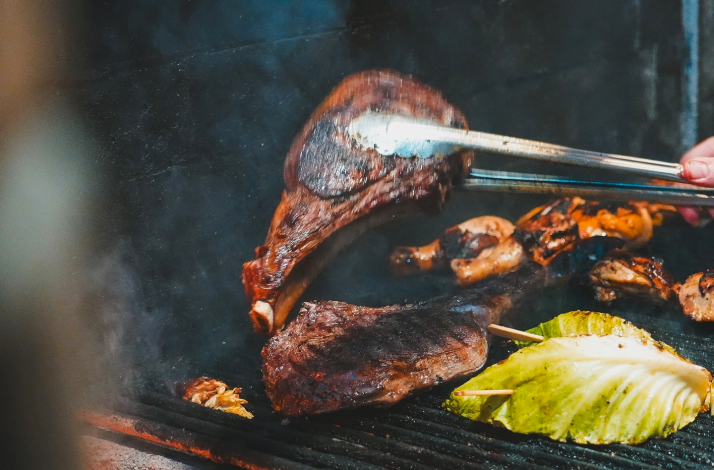 Large cuts of tomahawk steak and fresh greens being grilled over hot coals.