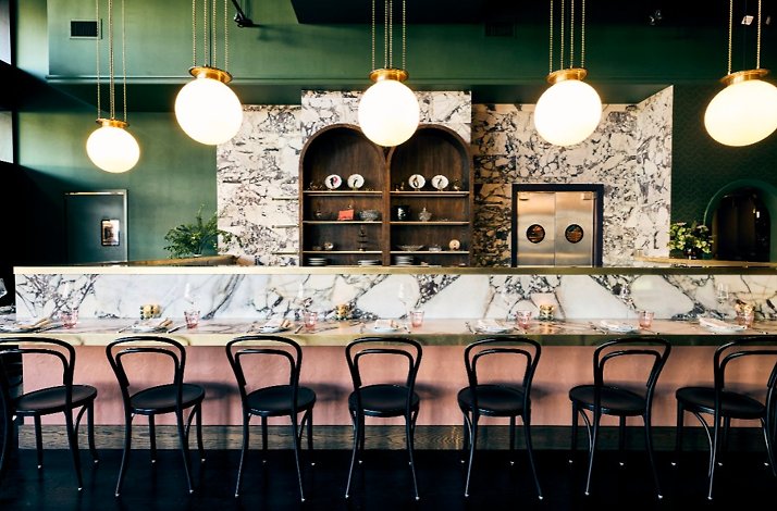 JouJou's marble bar counter lined with black bistro chairs, arched wood shelving, and globe pendant lights in a moody green-toned space.