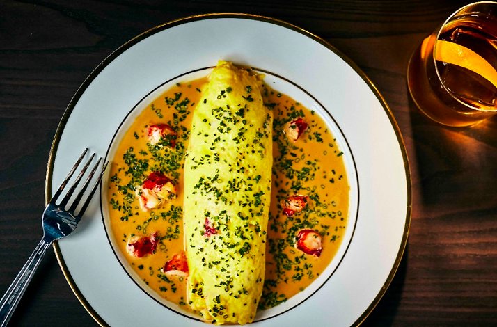 Rolled omelette in lobster cream sauce, topped with minced chives and surrounded by shellfish pieces, plated on a gold-rimmed white dish.