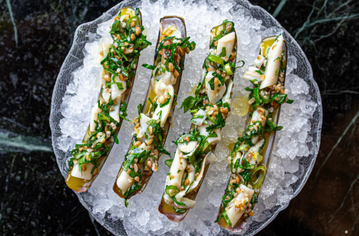 Four razor clam shells on crushed ice at Bar St-Denis, garnished with fresh herbs, grains, and green onion.