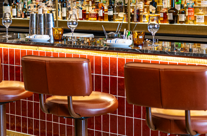 Bar St-Denis bar counter with brown leather stools, red-tiled facade, wine glasses, and a fully stocked liquor shelf backdrop.