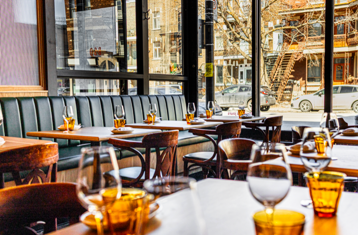 Sunlit interior of Bar St-Denis showing set dining tables with amber glasses and a view of a Montreal street outside.