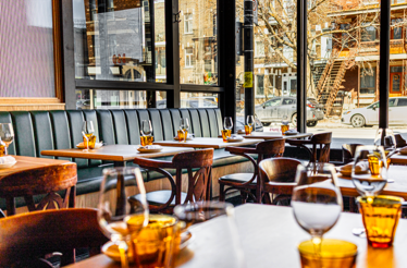 Sunlit interior of Bar St-Denis showing set dining tables with amber glasses and a view of a Montreal street outside.