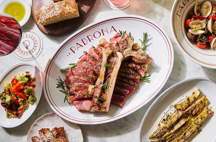 Sliced T-bone steak centred on a 'La Padrona'-branded oval platter, surrounded by clams, salad, focaccia, and grilled leeks.