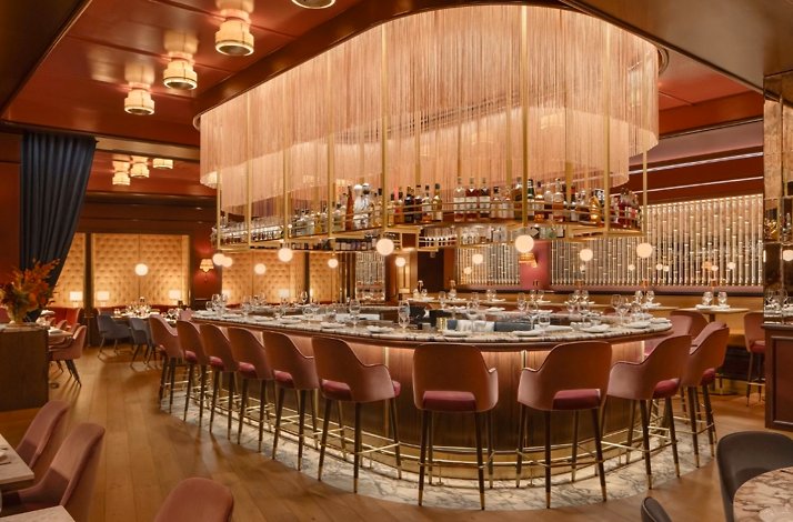 Oval bar at La Padrona Boston topped by a dramatic rose-gold fringe chandelier, surrounded by pink velvet bar stools.
