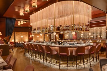 Oval bar at La Padrona Boston topped by a dramatic rose-gold fringe chandelier, surrounded by pink velvet bar stools.